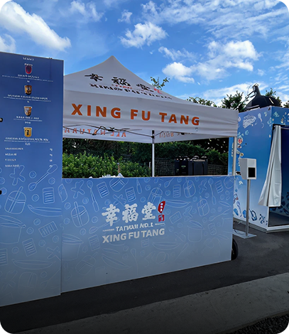 A booth with a blue counter displaying the logo and name of XING FU TANG, with a white canopy tent behind it bearing the same name. The booth has graphical elements resembling drinks and bubbles, and a blue sky with some clouds is visible above.