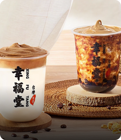 Two cups of bubble tea with brown and white swirls on top, one with a white cup on a wooden coaster, the other on a round wooden slab, with coffee beans and cornflakes nearby.