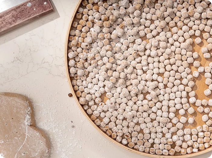 Close-up of a bamboo tray filled with small beige and gray dried chickpeas or split peas, with a flour-dusted surface nearby.