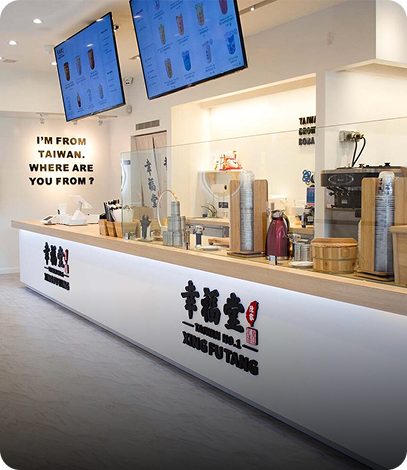 Inside a modern bubble tea shop with a white counter and two large digital menu screens, featuring beverages, and signage indicating it is from Taiwan, with the shop name 'Xing Fu Tang' in both English and Chinese.