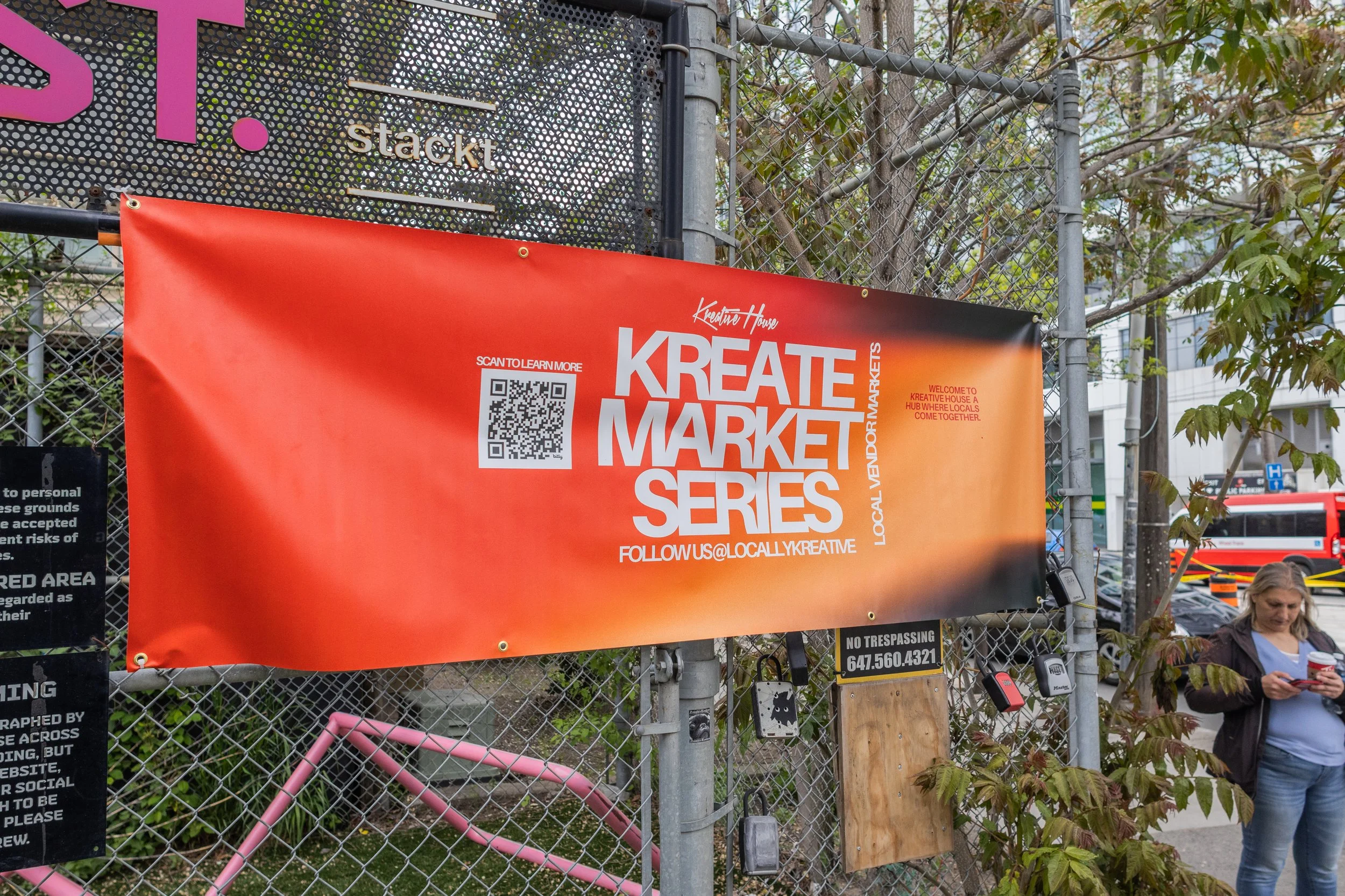 Pink and orange banner promoting the 'Kreate Market Series' event, with a QR code, social media handle, and an area for vendors, attached to a chain-link fence near trees and a woman looking at her phone.