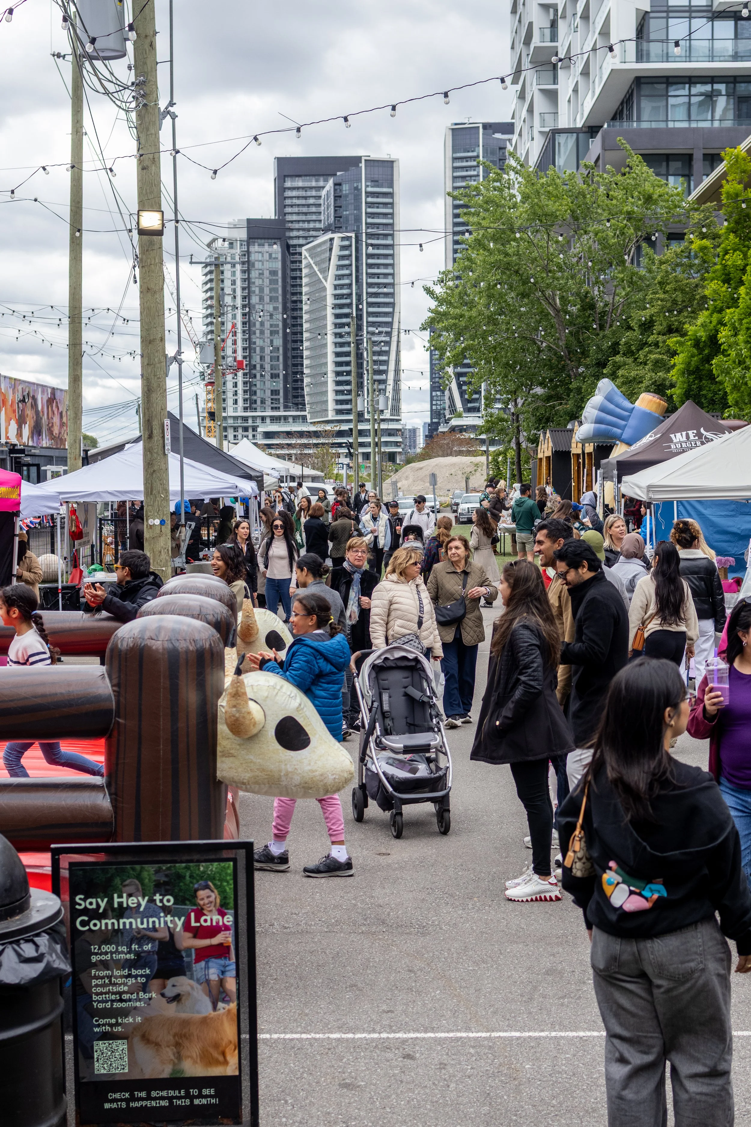 A busy outdoor street market with people walking and shopping under gray cloudy skies. There are vendor booths on both sides, a large inflatable cow, and tall modern buildings in the background.