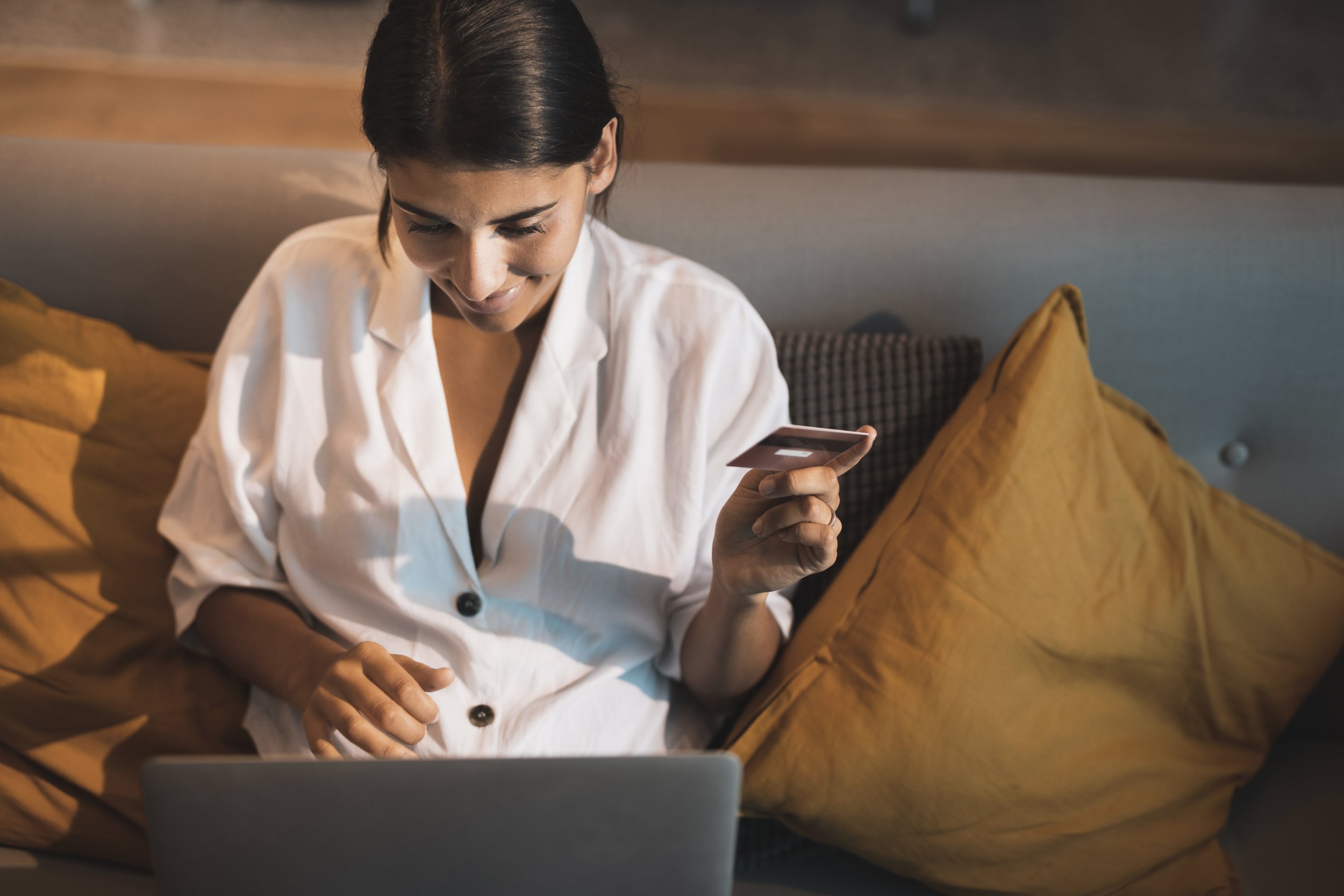 Woman sitting on a couch with yellow pillows, using a laptop and holding a credit card, smiling while shopping online.