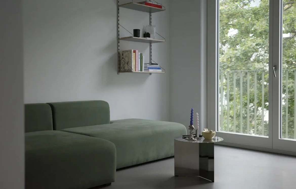 Living room with green sectional sofa, small round coffee table holding candles and a bowl, white wall with a wall-mounted shelf holding books and decor, and a sliding glass door leading to a balcony with green trees outside.