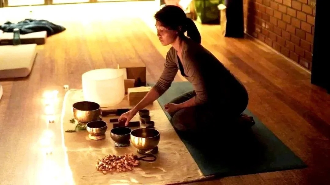 A woman practicing sound healing with singing bowls on a wooden table in a dimly lit room.