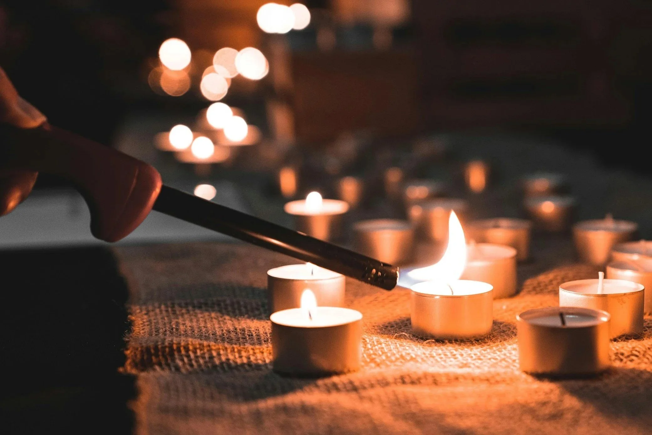 A hand holding a lit matchstick igniting a tealight candle among several lit tealights on a textured surface in a dark environment.
