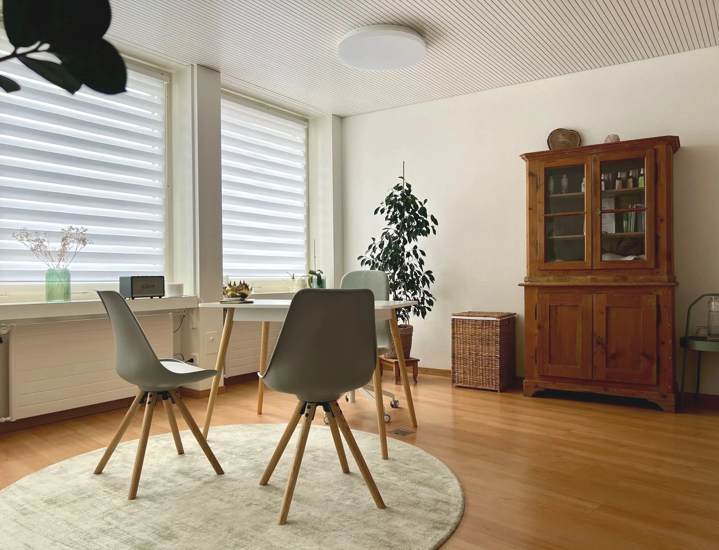Heller, minimalistischer Essbereich mit vier Stühlen um einen weißen Tisch, große Fenstervorhänge, eine grüne Vase mit Trockenblumen, eine große grüne Zimmerpflanze, eine alte Holzkastenschrank, ein geflochtener Korb, ein kleiner Beistelltisch und ein runder Teppich auf Holzboden.