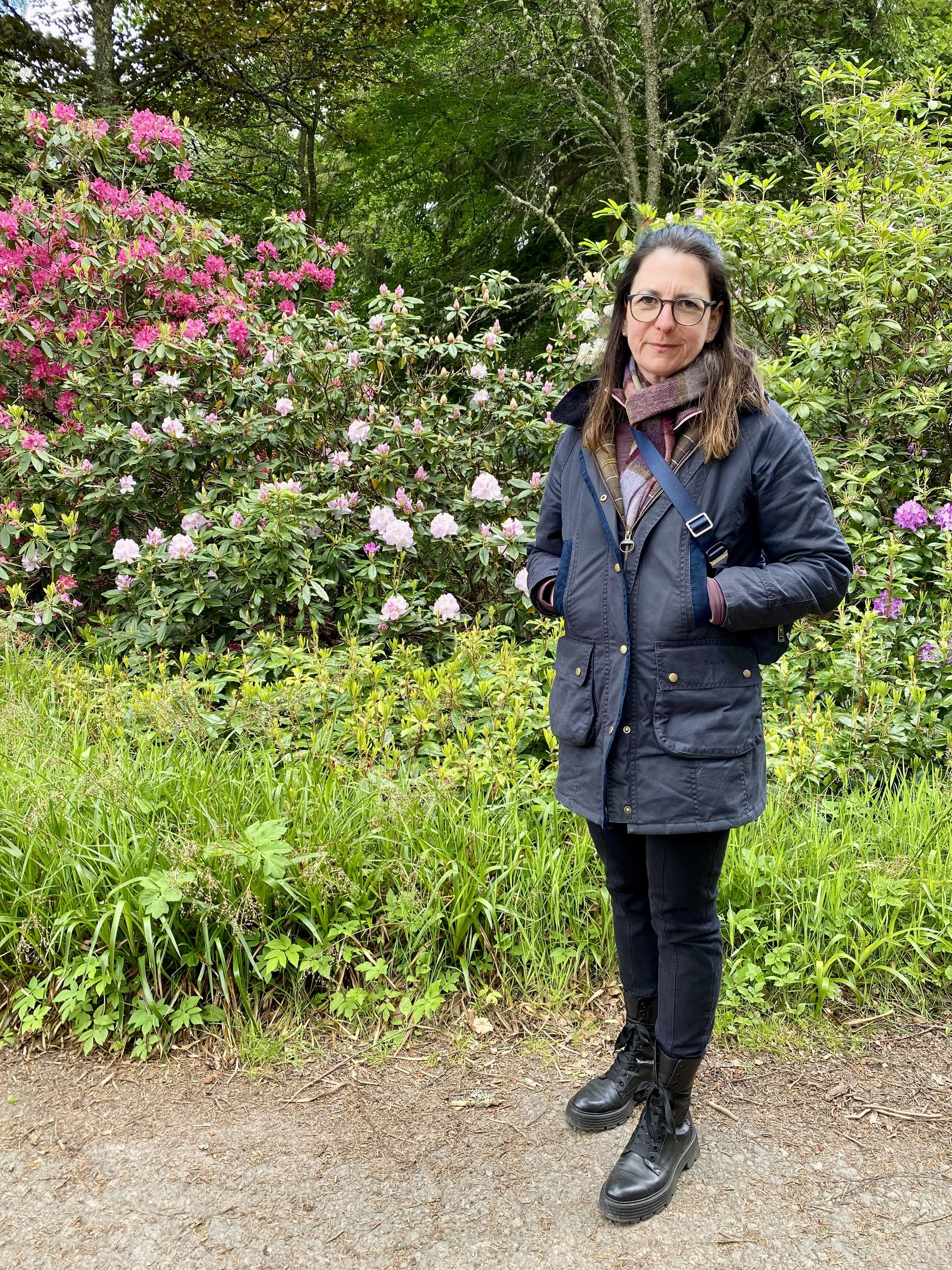 Frau steht vor blühenden Rhododendron im Garten bei bewölktem Himmel, trägt dunkle Jacke, schwarze Hose, Wanderstiefel und Brille.