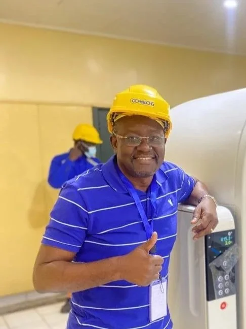 A smiling man wearing a yellow hard hat and blue striped polo shirt giving a thumbs-up, with another person in the background also wearing a yellow hard hat, in an indoor setting next to a water dispenser.