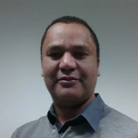 Portrait of a man with short dark hair, wearing a gray collared shirt, standing against a plain light gray wall.