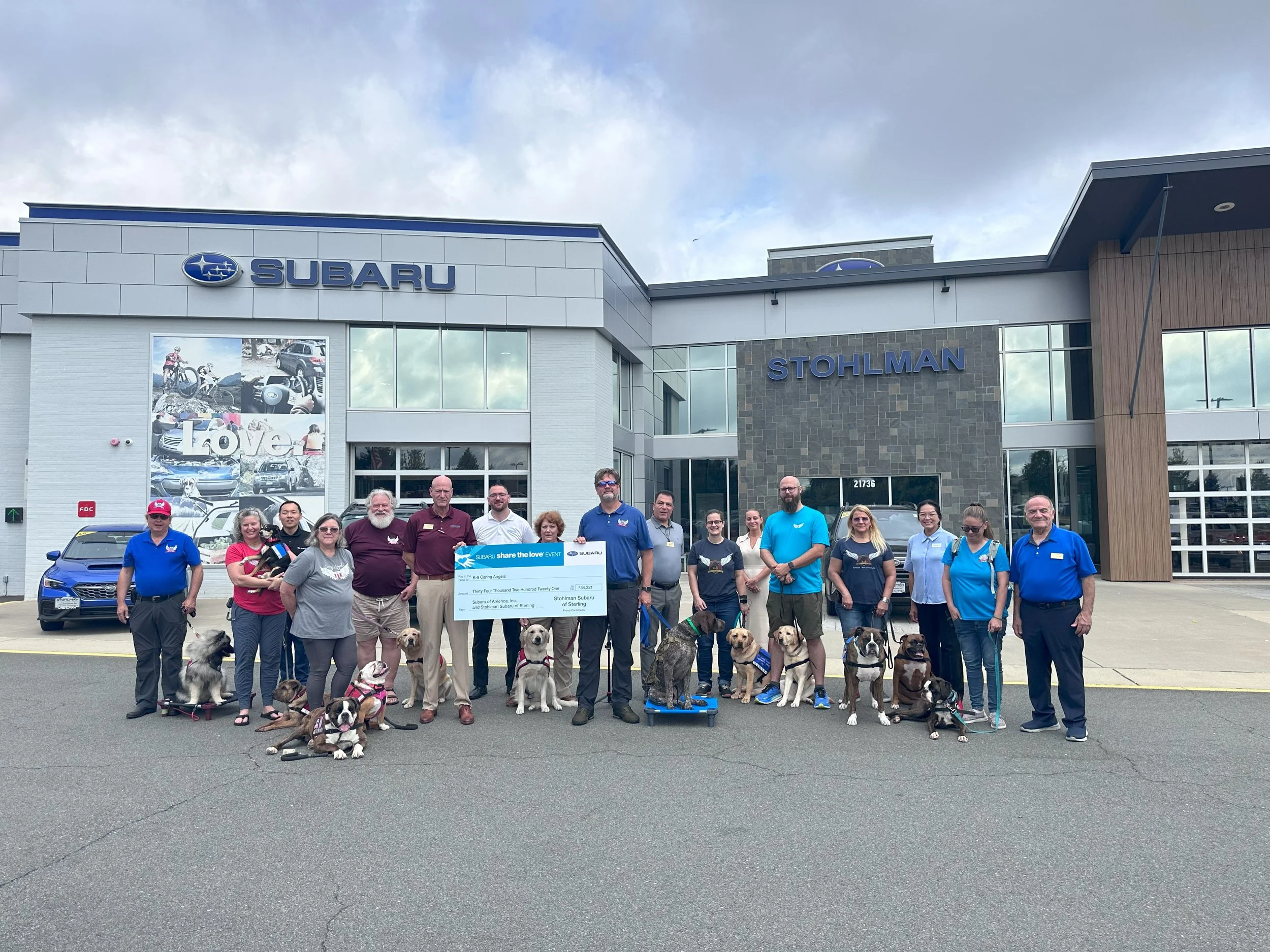 Group of people and dogs standing in front of Subaru dealership holding a large check for a charity event.