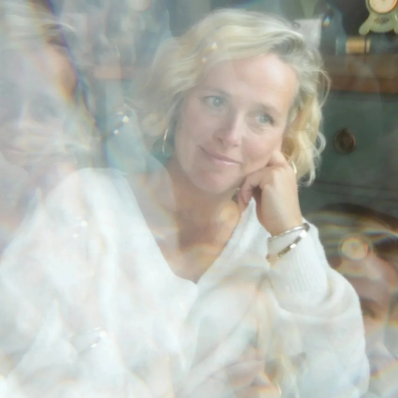 A woman with blonde curly hair resting her chin on her hand, wearing a white sweater and jewelry, sitting in a room with a blurred background.