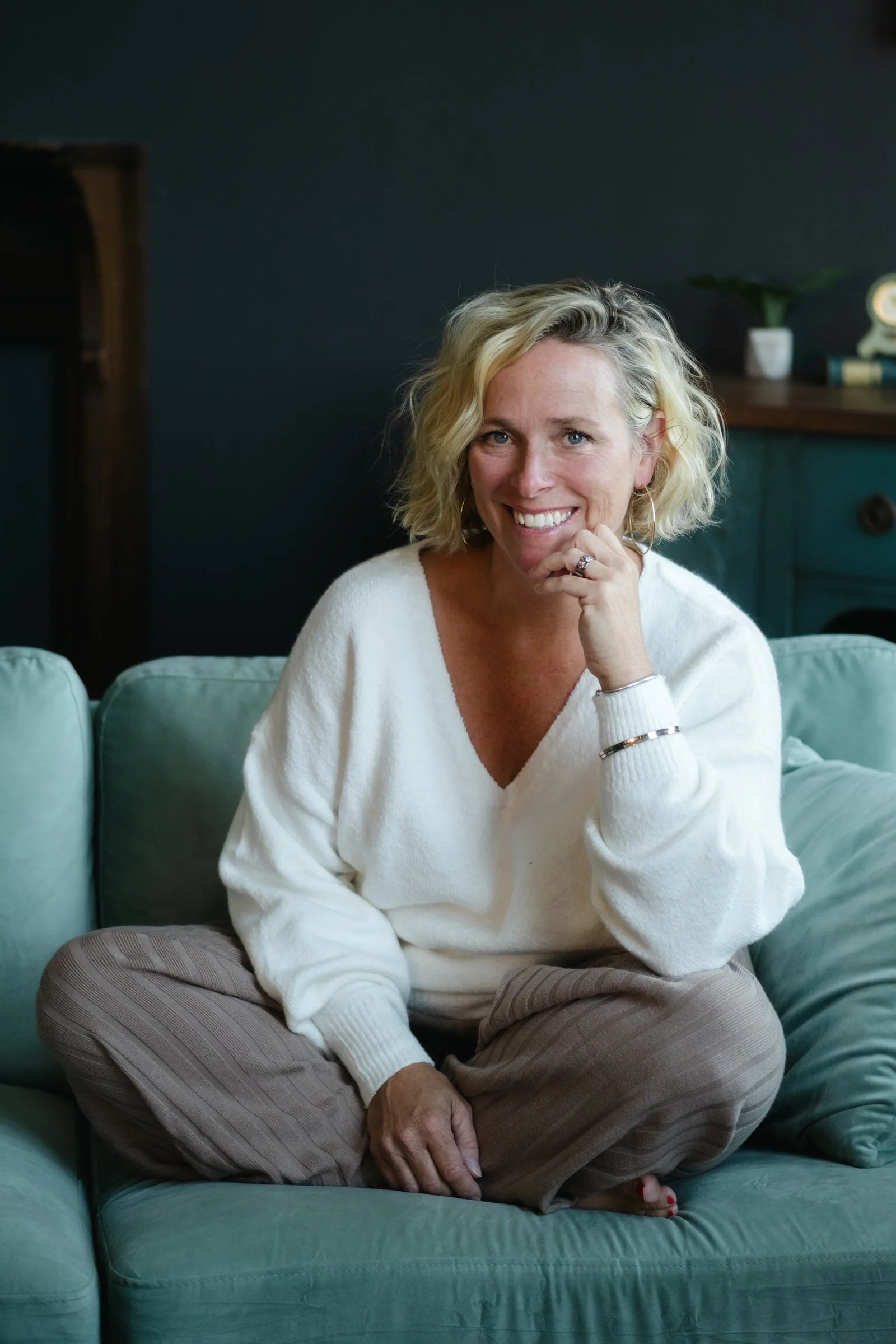 A woman with blonde, curly hair sitting cross-legged on a teal sofa, smiling, with a dark wall and side table with decor in the background.
