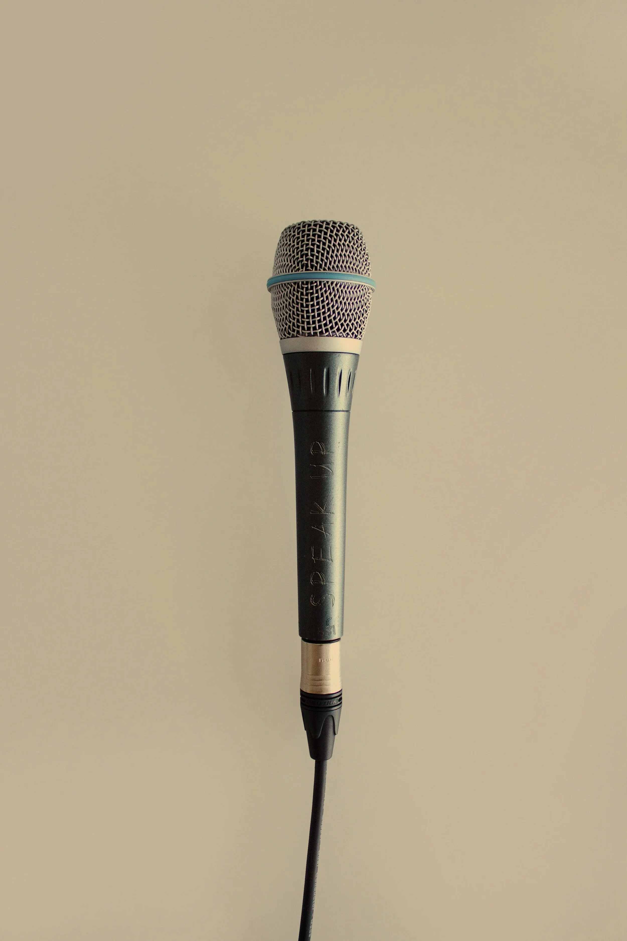A close-up of a microphone with a black handle and a metallic mesh head, hanging against a plain beige wall.