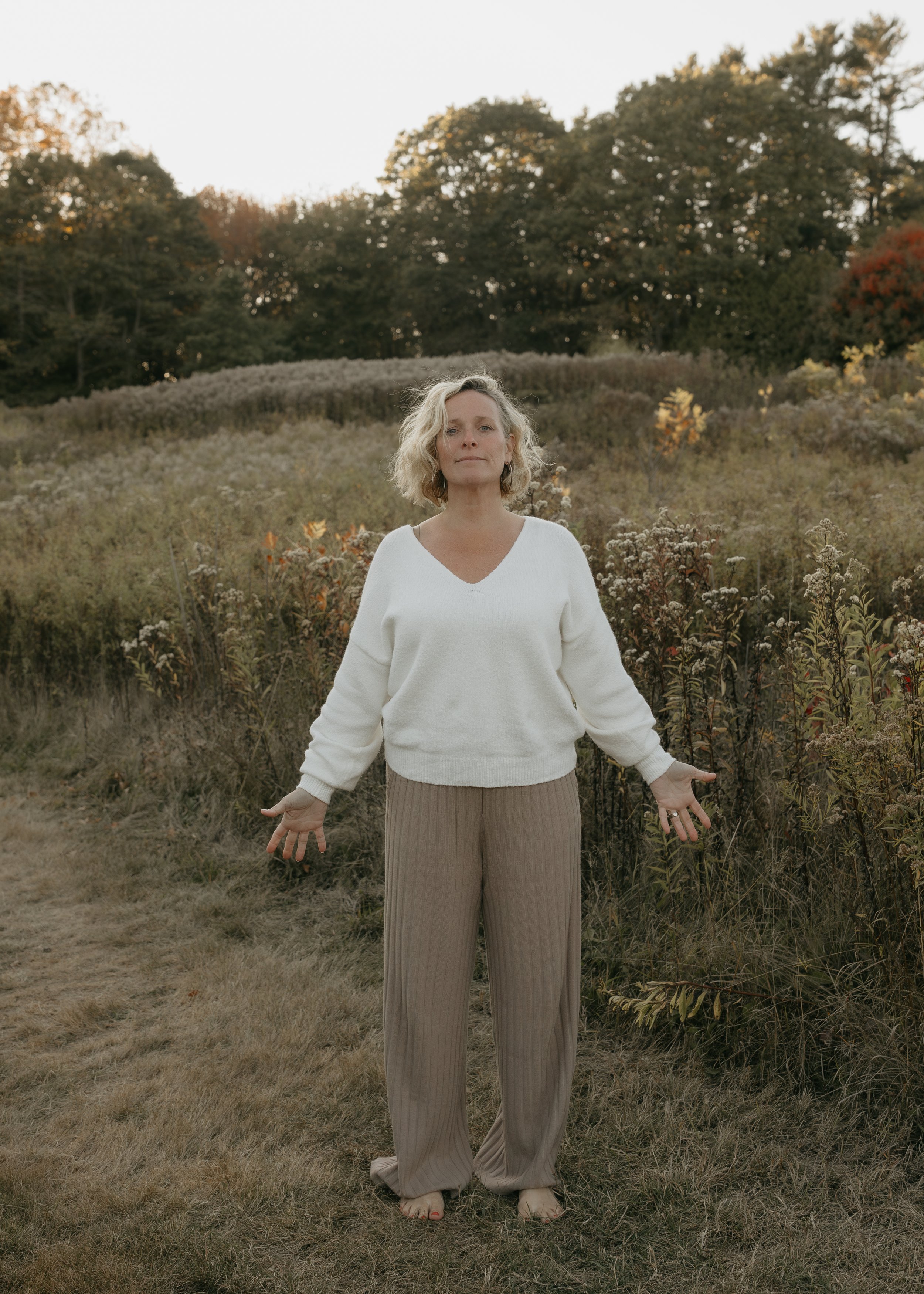 A woman standing outdoors in a field with trees in the background during fall, wearing a white sweater and beige pants, with her arms outstretched and a peaceful expression.