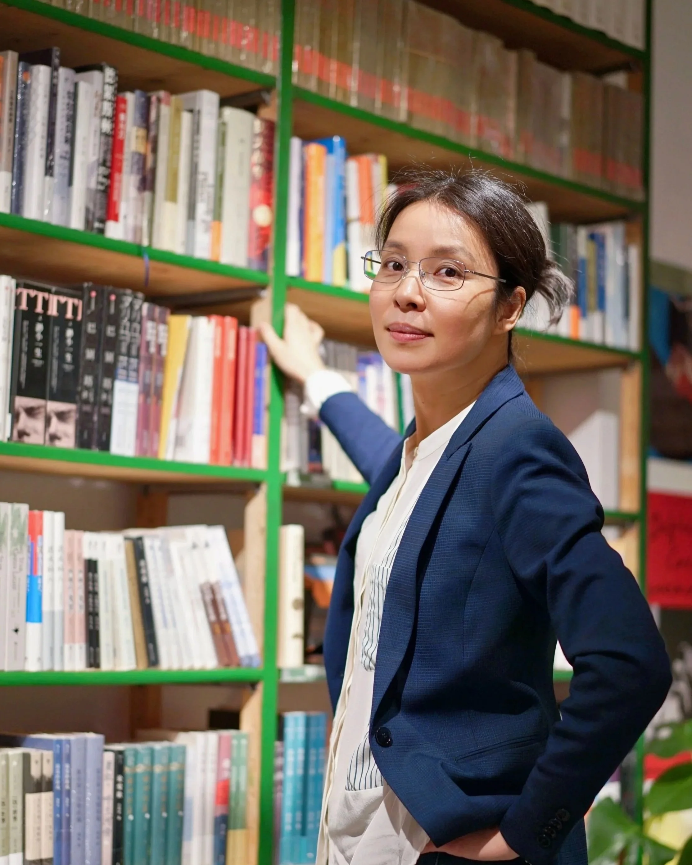 A woman in a blue blazer and white shirt standing in front of bookshelves in a library, reaching for a book on the shelf.