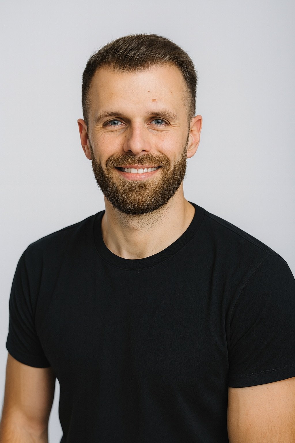 Portret van een lachende man met een baard en korte haar, gekleed in een zwarte T-shirt, tegen een effen lichte achtergrond.