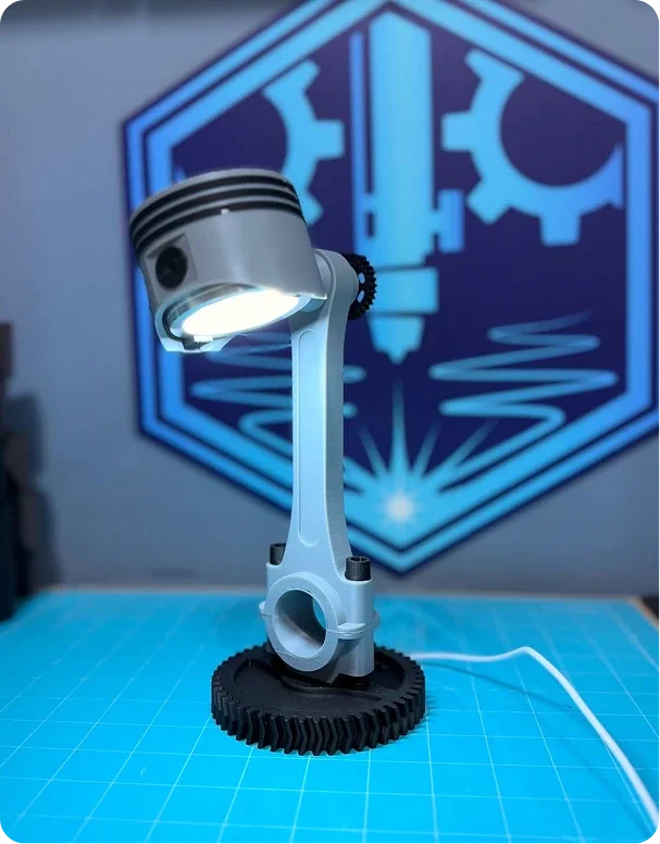 A small, desktop lamp designed to look like a piston and crankshaft, with a gear at the base and a bright LED light, set on a blue cutting mat with a tech-inspired logo on the wall behind.
