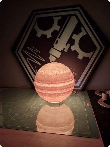 Decorative spherical lamp resembling a planet, placed on a grid-patterned surface, with a gear and laboratory equipment silhouette in the background.