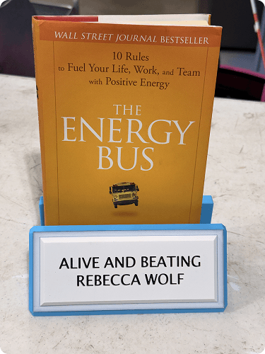 A book titled 'The Energy Bus' by Jon Gordon placed in a blue holder on a table, with a sign in front that reads 'Alive and Beating Rebecca Wolf'.