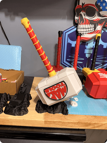 A Mjolnir hammer toy with a yellow handle and red accents, a red toy hammer, and a skull decoration mounted on a wall in the background.
