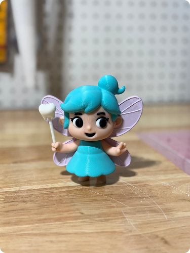 Toy figurine of a fairy with blue hair, purple wings, and a blue dress, holding a wand with a heart-shaped top, placed on a wooden surface.