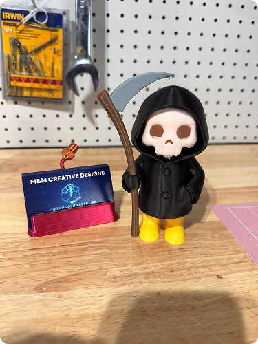 A small figurine with a skull face, wearing a black hooded cloak and yellow boots, holding a scythe, placed on a wooden surface with tools and a pegboard in the background.
