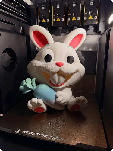 A plush toy rabbit with large eyes, a pink nose, and big front teeth, sitting inside a 3D printer or similar machine.