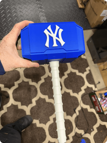 A person holding a blue plastic tool with the New York Yankees logo on it. The tool has a white handle. The background features a brown patterned rug and some boxes.