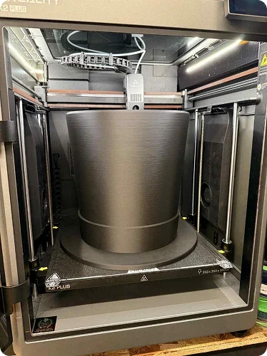 A 3D printer with a large, cylindrical object being printed inside the enclosed printer chamber.