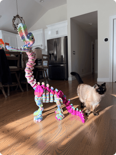 Colorful, coiled decoration resembling a dinosaur sculpture on a hardwood floor, with a Siamese cat nearby in a home interior.