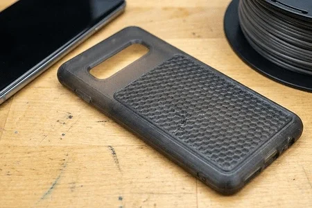 Black rubber phone case on a wooden desk next to a partial view of a wireless charger and some electronic devices 3d printed using TPU filament.