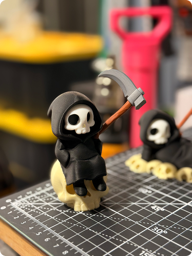 Small Halloween figurine of a skeleton with a hood, holding a tiny pickaxe, standing on a resin skull, with other similar figures in the background on a craft cutting mat.