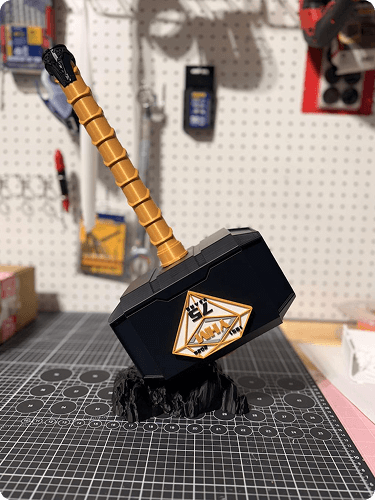 A miniature replica of Thor's hammer, Mjolnir, with a gold handle and black head, displayed on a black textured base in a workshop or store setting.