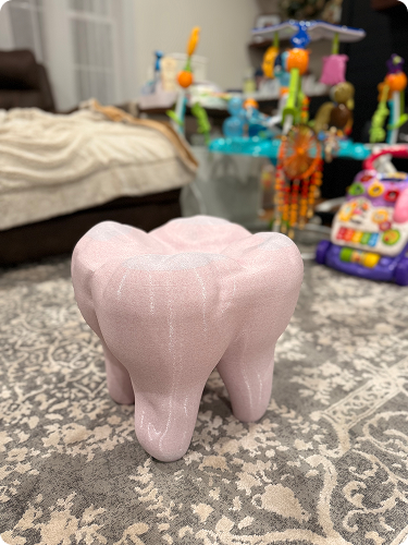 Pink, textured toy in the shape of a tooth on a patterned rug in a living room with a play area and toys in the background.