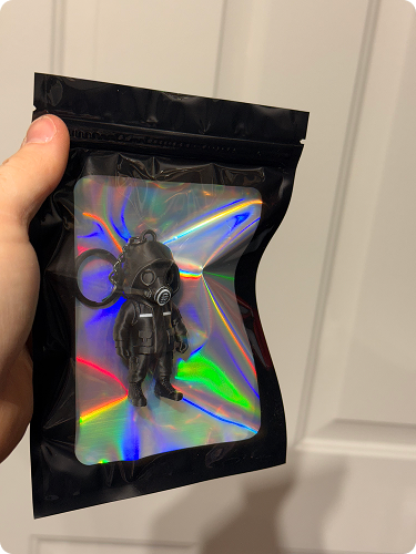 Person holding a holographic plastic bag containing a black keychain with a figure wearing a gas mask and a suit.