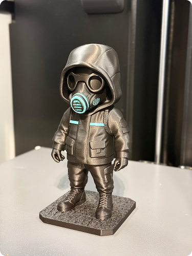 A small figurine of a person wearing a hooded jacket, pants, boots, glasses, and a gas mask, standing on a rectangular base.