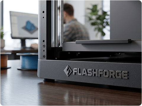 Close-up of a FlashForge 3D printer on a wooden table, with a person working at a computer in the background.