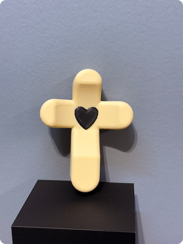 Yellow cross-shaped figure with a black heart in the center, placed on a black pedestal against a gray wall.