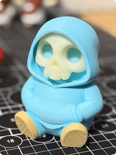 A blue figurine with a skull face inside a hoodie, sitting on a black surface with white markings.