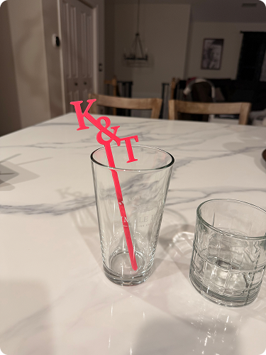 A tall clear glass with a pink K and T straw inside on a white marble table, next to a shorter glass with a textured pattern.