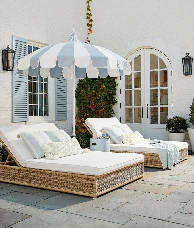 Outdoor patio with two wicker lounge chairs with white cushions, a large blue and white striped patio umbrella, a small white side table, potted plants, and a white brick house with blue shutters in the background.