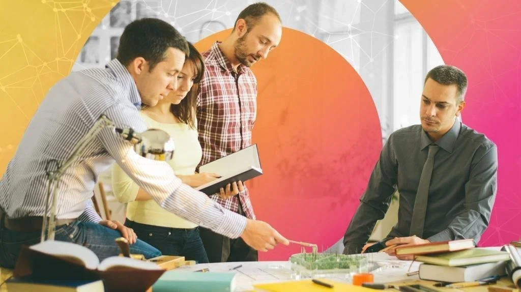 Group of four professionals discussing a project at a table with books and a model of greenery, in a modern office with digital artwork in the background.