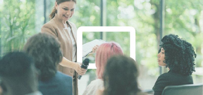 Workshops: 
Build the Capabilities Complex Challenges Require [A woman holding a microphone and a clipboard presenting to a group of diverse people in a well-lit, green indoor space.]