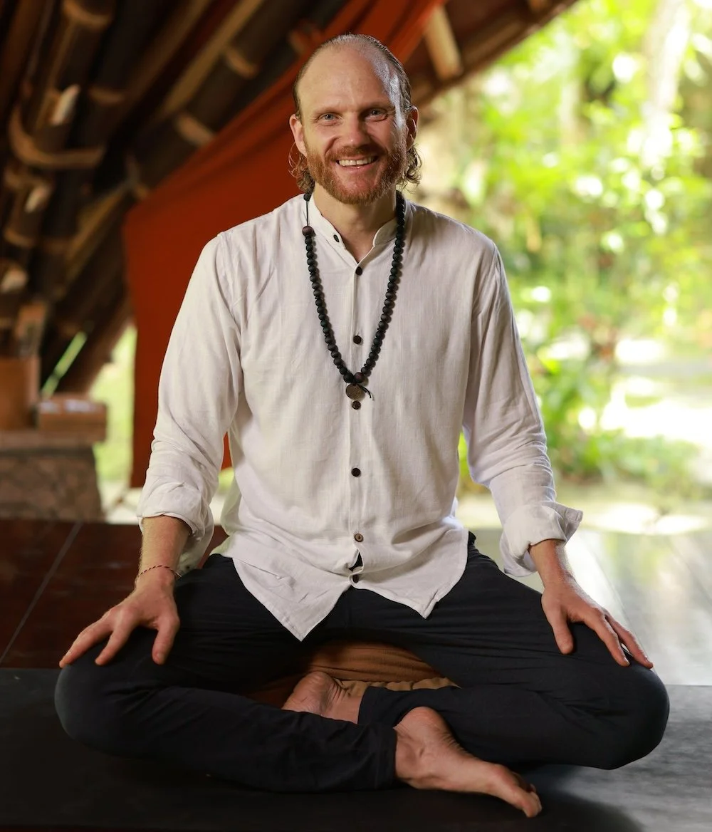 A man with a beard and long hair sitting cross-legged on the floor, smiling, wearing a white shirt and black pants with a beaded necklace, in a room with wooden beams and greenery outside.