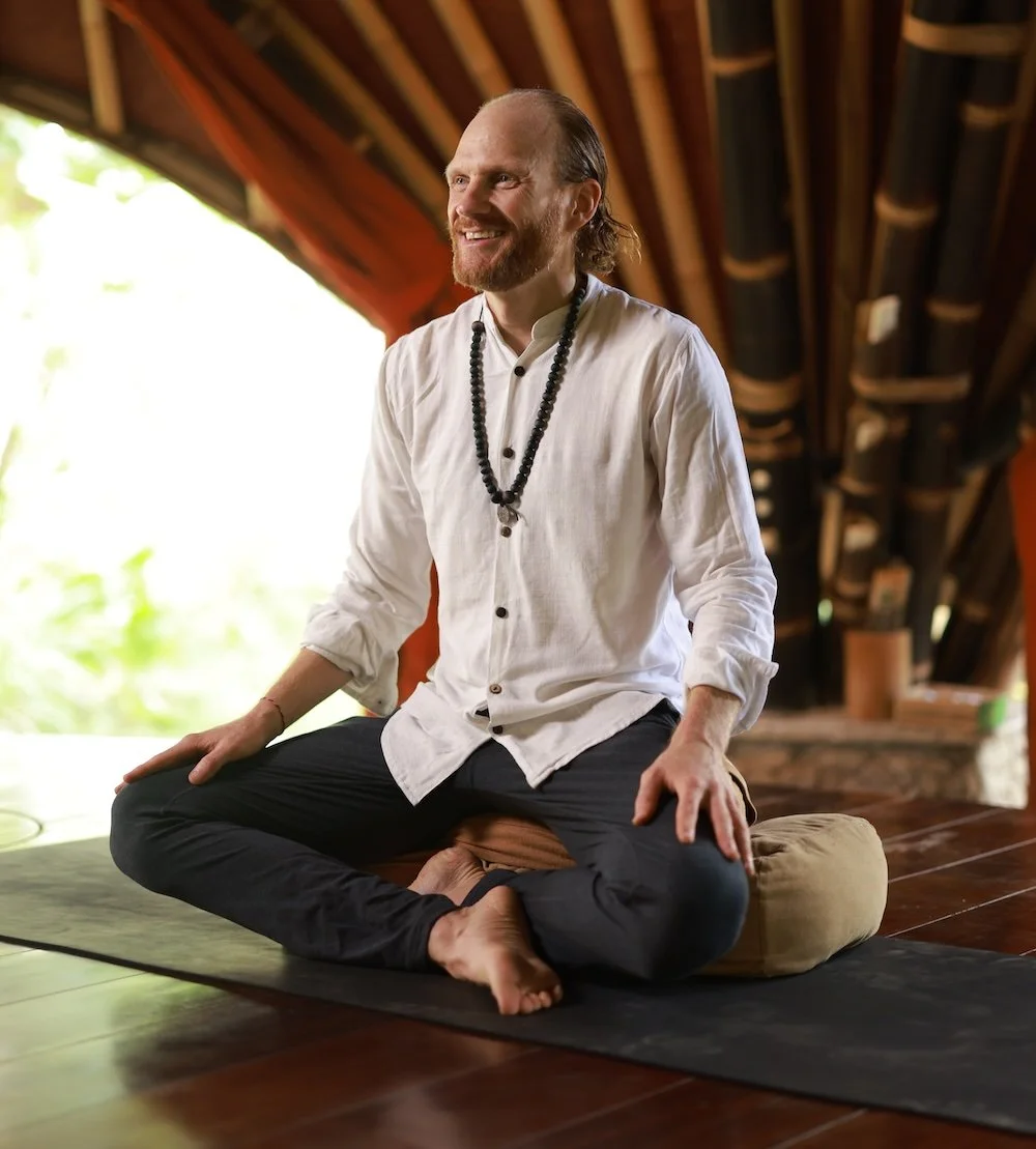A man with a beard and long hair, smiling and sitting cross-legged on a cushion on a yoga mat, inside a wooden-floored room with wooden beams. He is wearing a white shirt and a beaded necklace, with a background of greenery outside.