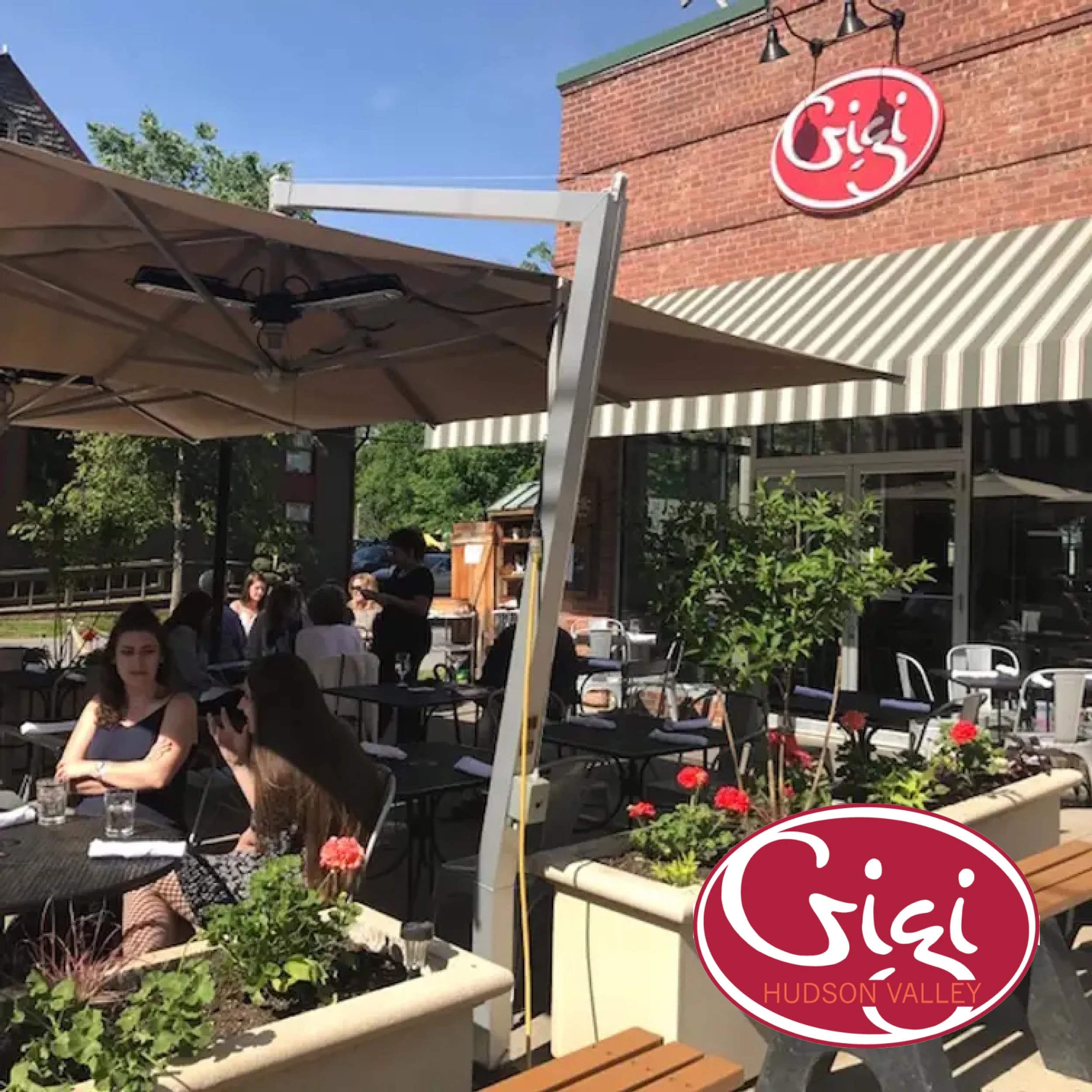 Outdoor patio of Gigi restaurant in Hudson Valley with groups of people dining, large umbrellas, a brick building, planters with flowers, and string lights.