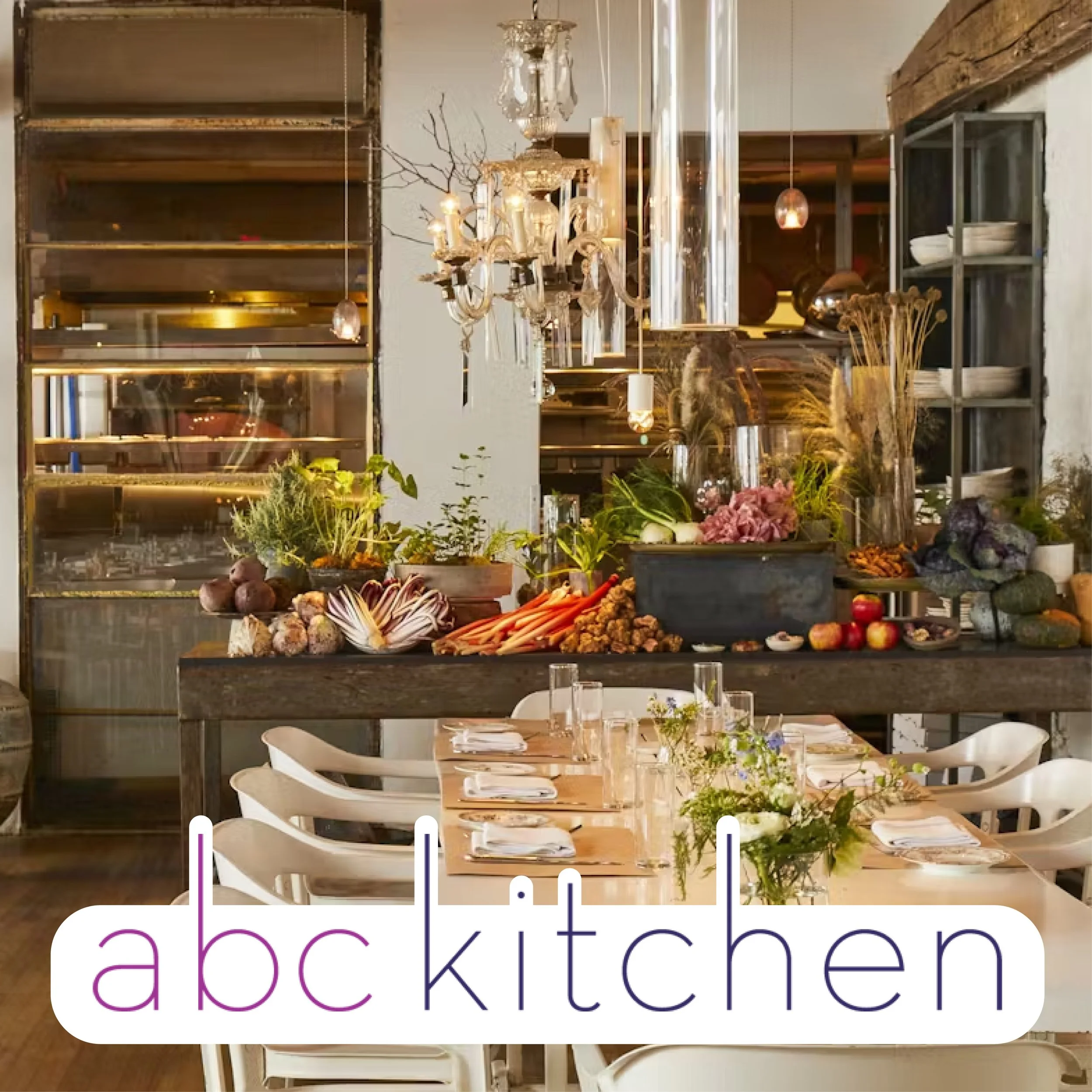 A dining table set for a meal with a floral centerpiece, surrounded by white chairs, and a background featuring a rustic interior with a chandelier and an abundance of fresh produce and plants on a wooden shelf.