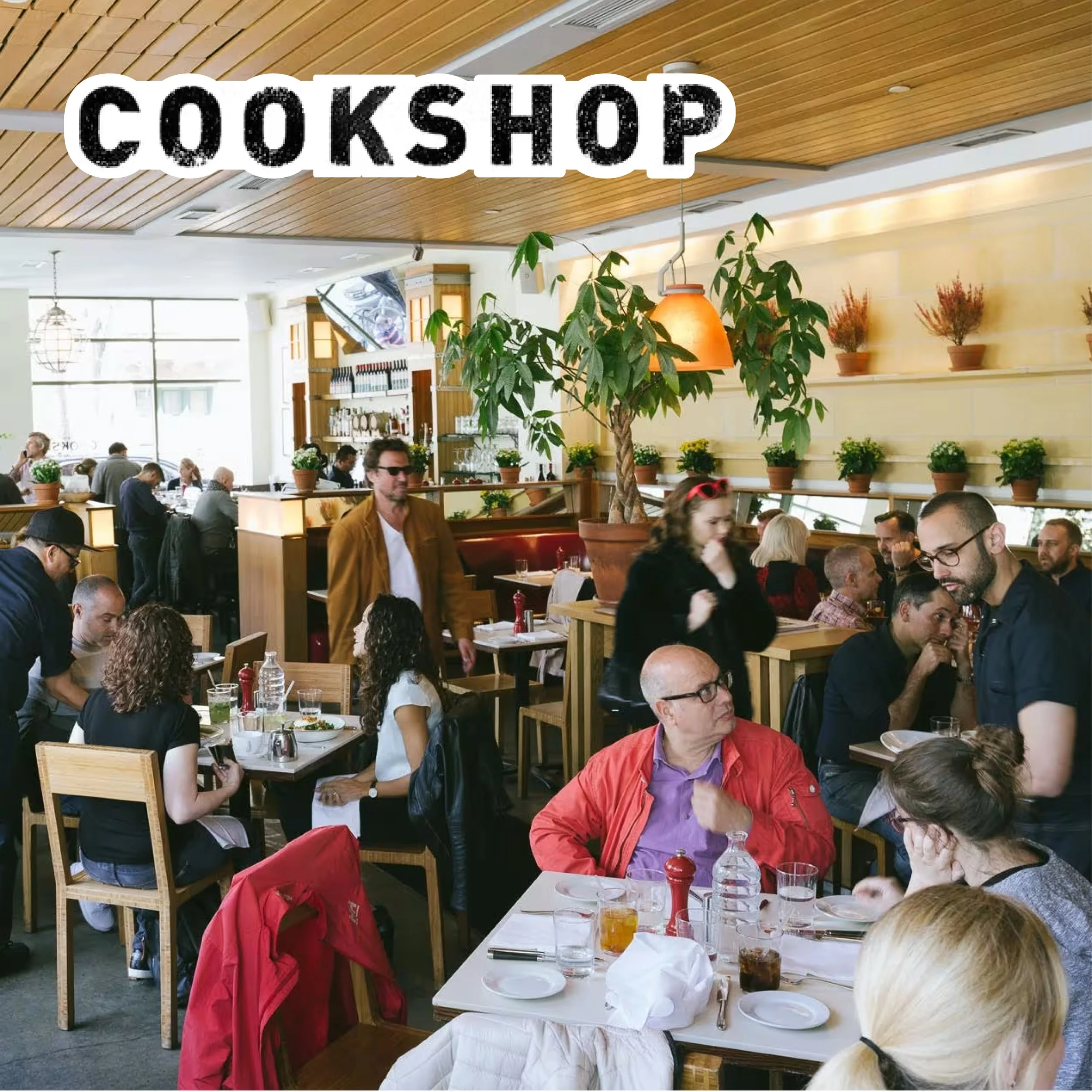 A busy restaurant with people dining and interacting, decorated with potted plants and warm lighting.