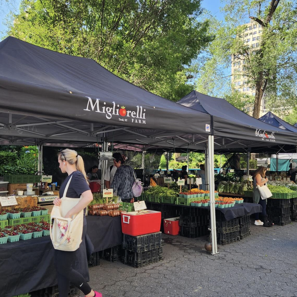 Outdoor farmers market booth under a black canopy with the sign "Migliorelli FARM". There are fresh vegetables and herbs displayed on tables. People are shopping, and trees and a tall building are visible in the background.